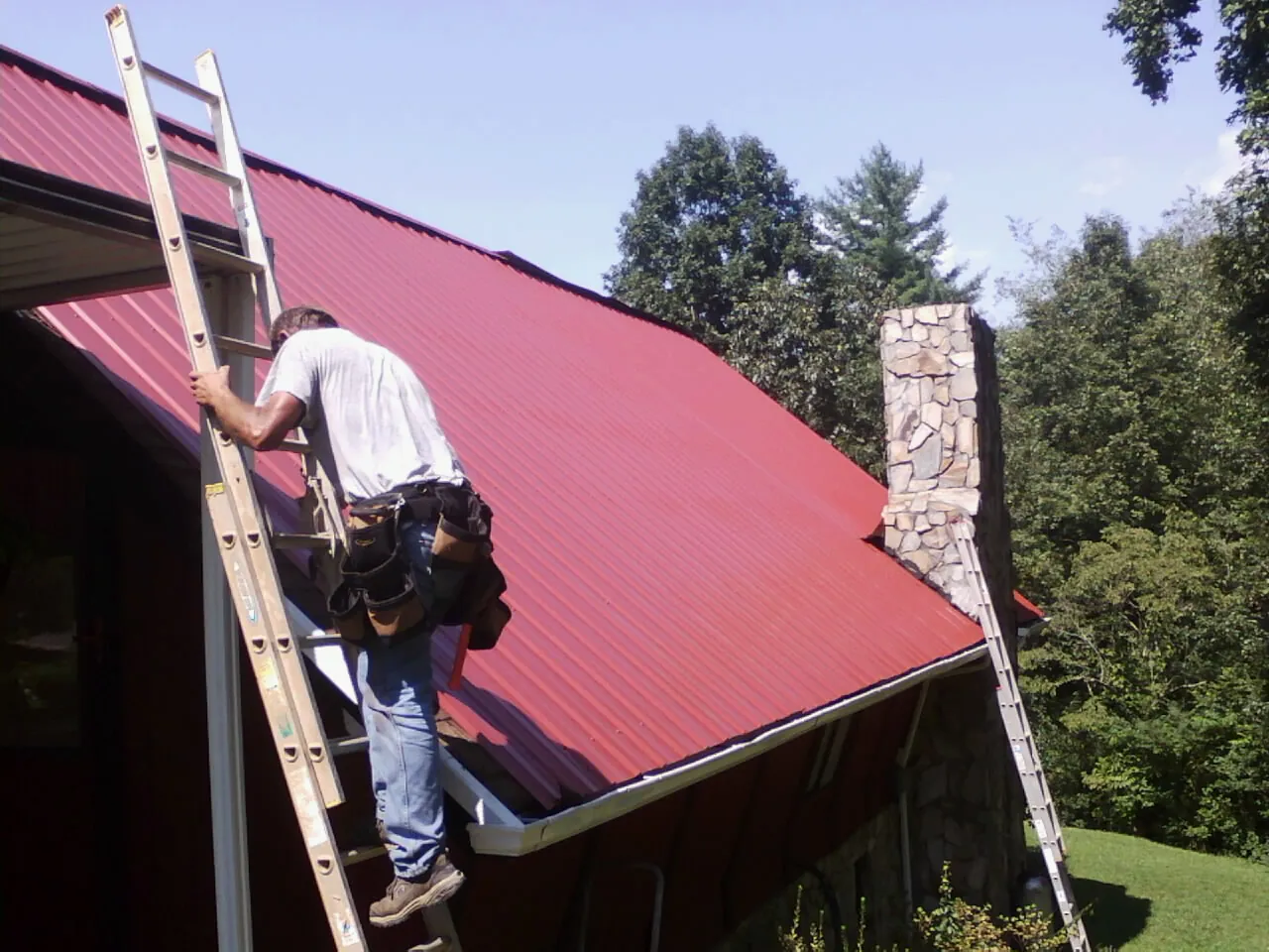 Roofer on a ladder beside a red metal roof and stone chimney — Soffit & Fascia Repair by Roofing Brothers in Rockaway Beach